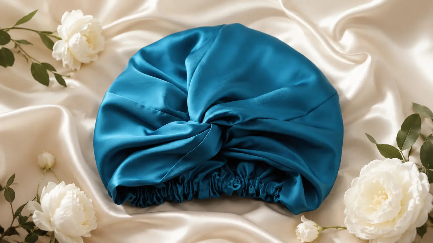 Bonnet en soie bleu pétrole posé sur satin champagne avec fleurs blanches et feuillage délicat