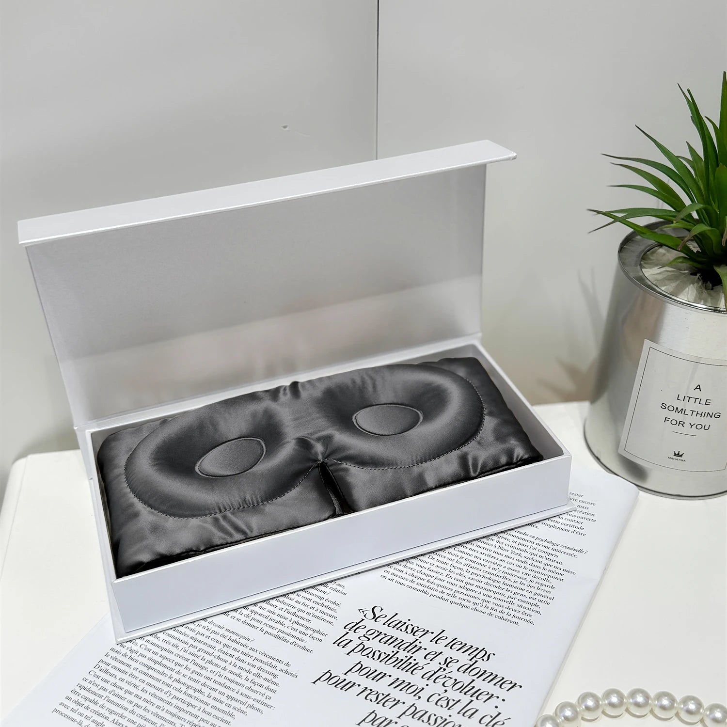 Masque en soie gris anthracite, posé dans une boîte blanche, entouré de décorations élégantes et d'une plante verte