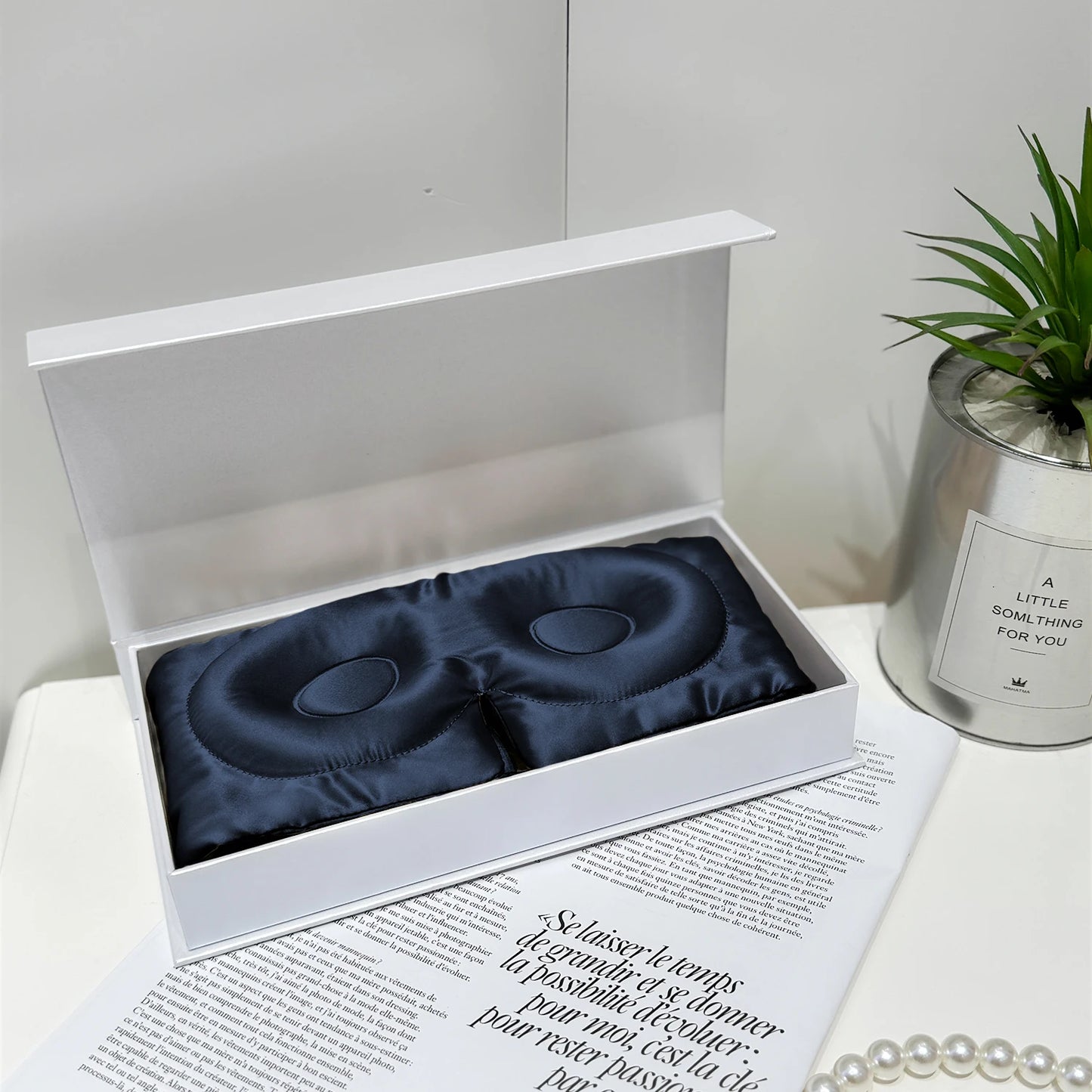 Masque de sommeil en soie bleu marine, présenté dans une boîte blanche, sur une surface décorée de magazines et d'une