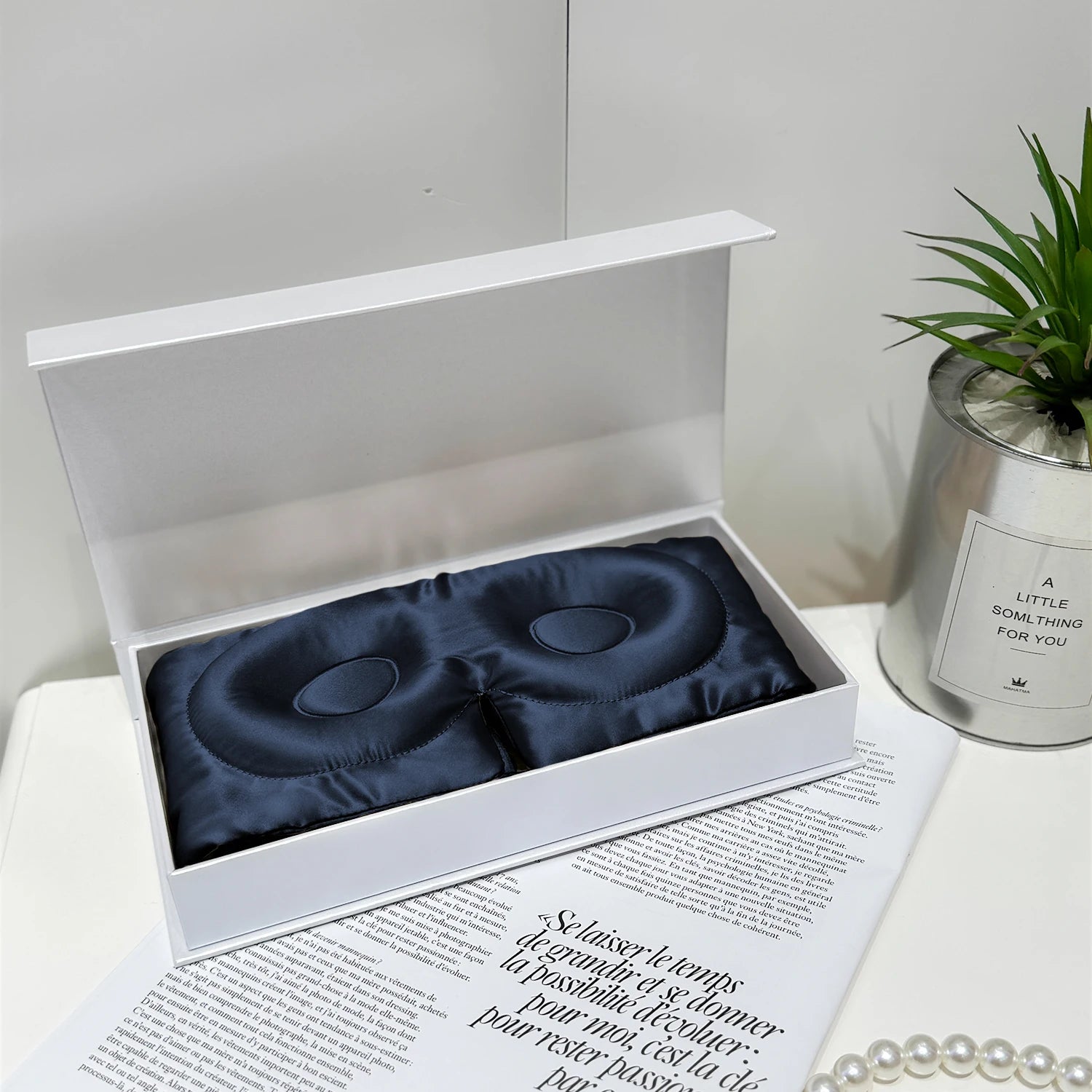Masque de sommeil en soie bleu marine, présenté dans une boîte blanche, sur une surface décorée de magazines et d'une