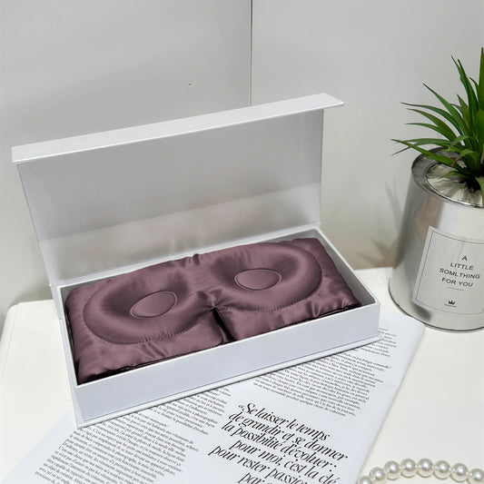 Masque de sommeil en soie mauve grise, délicatement posé dans une boîte blanche sur un fond neutre
